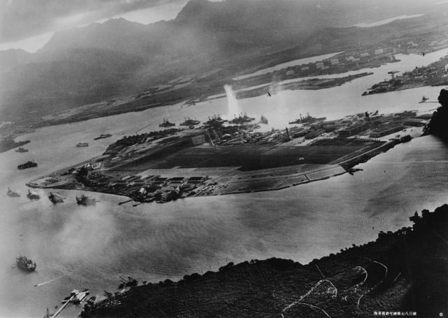Photograph of Battleship Row taken from a Japanese plane at the beginning of the attack. The explosion in the center is a torpedo strike on USS West Virginia. Two attacking Japanese planes can be seen: one over USS Neosho and one over the Naval Yard.