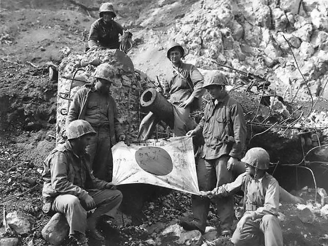 U.S. Marines pose on top of enemy pillbox with a captured Japanese flag.
