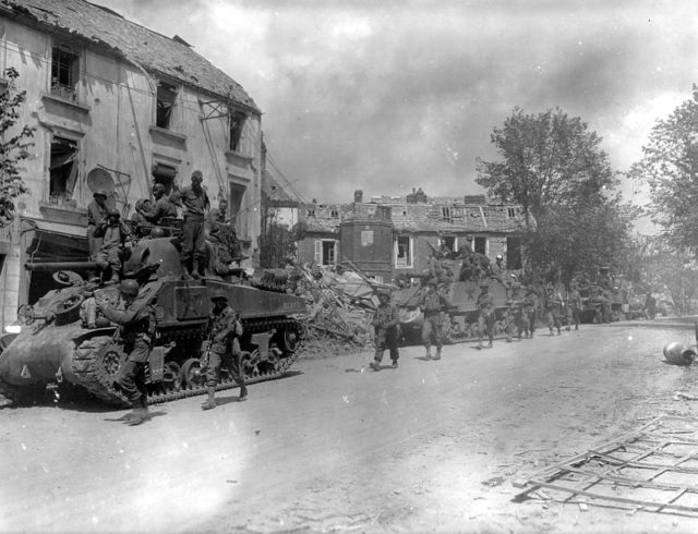 American armored and infantry forces pass through the battered town of Coutances, France, during Operation Cobra. By Unknown - US National Archives - 111-SC-199774, Public Domain, https://commons.wikimedia.org/w/index.php?curid=6495321