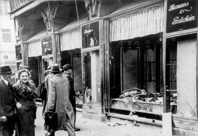 Germans smile while walking past a Jewish shop damaged on Kristallnacht, Magdeburg, 1938. Photo Credit.