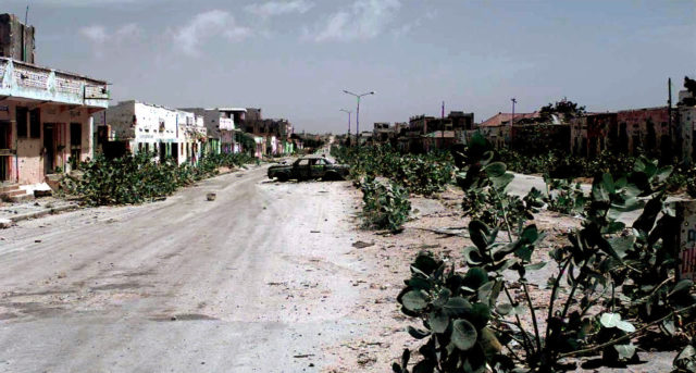 A long shot an abandoned Mogadishu Street known as the "Green Line". Foliage has grown up along the sidewalk on both sides of the street. An abandoned, burned out car is seen in the center of the frame. The street is the dividing line between North and South Mogadishu, and the warring clans. Members of the clans (not shown) tore down the roadblocks along the line in a show of unity. This mission is in direct support of Operation Restore Hope.
