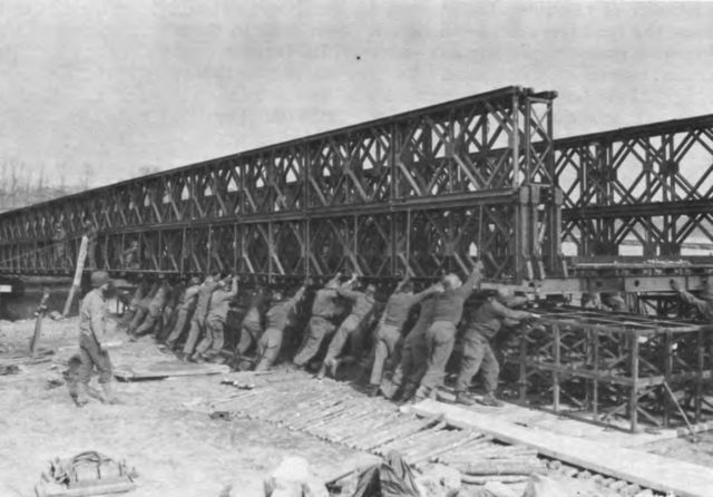 Engineers slide a Bailey Bridge section into place, almost every part of the bridge construction was done by hand. The only time heavy equipment was used was to lift pieces into high places. Image Source: 