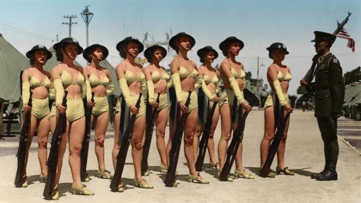 August 01, 1935 An unidentified corporal from the US Army’s 30th Infantry Division (based in the San Francisco Presidio) stands at attention with a group of women in two-piece bathing costumes, doughboy helmets, and heels, each of whom holds an M1917 Enfield rifle, on a parade ground in Camp George Derby, on the grounds of the California Pacific International Exposition in Balboa Park, San Diego California, mid to late 1935. Mostly obscured behind the women is the entrance to the expo’s so-called ‘Indian Village.’ Paul Reynolds / mediadrumworld.com