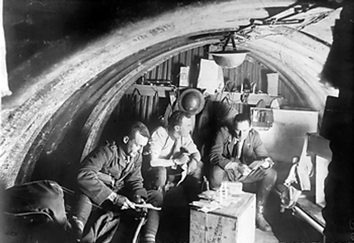 Interior of a dugout occupied by officers of the Australian 105th Howitzer Battery of the 4th Brigade. Three officers are looking at papers in the light of two candles on an upturned box.
