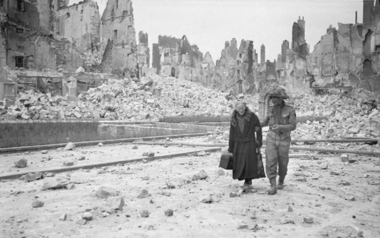 A British soldier in Caen after its liberation, gives a helping hand to an old lady amongst the scene of utter devastation.