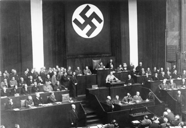 Adolf Hitler addressing the Reichstag on 23 March 1933. Seeking assent to the Enabling Act, Hitler offered the possibility of friendly co-operation, promising not to threaten the Reichstag, the President, the States or the Churches if granted the emergency powers. Photo Credit.
