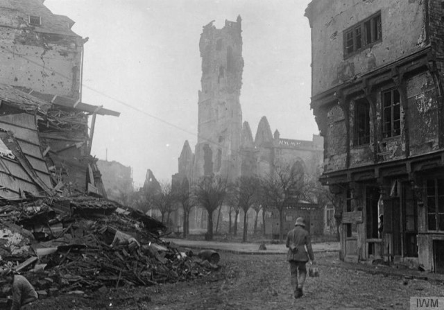 German soldier with a telephone wire drum in shell ruined and deserted city. July, 1916.