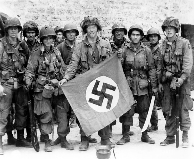American paratrooper, among the first to make successful landings on the continent, holds a Nazi flag captured in a village assault. Utah Beach, St. Marcouf, France. 8 June 1944 (Image).