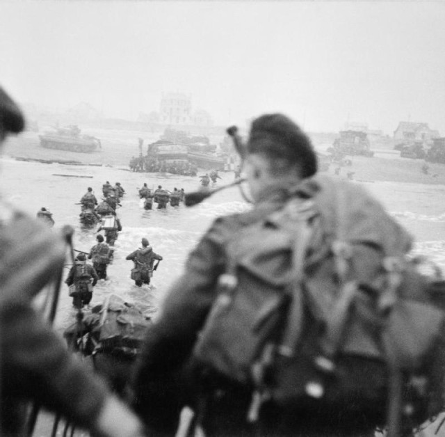 Sword Beach. On the right of the column, wades through the water. The figure in the foreground is Piper Bill Millin. By Evans, J L (Capt), No 5 Army Film & Photographic Unit - This is photograph B 5103 from the collections of the Imperial War Museums (collection no. 4700-29), Public Domain.