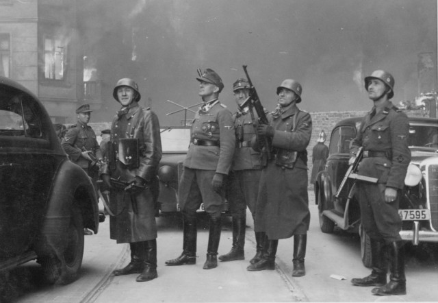 Stroop Report original caption: "The leader of the grand operation." SS-Brigadeführer Jürgen Stroop (center) watches housing blocks burn. The SD-Rottenführer at right is Josef Blösche ("Frankenstein"). Photo taken at Nowolipie street looking east, near the intersection with Smocza street. On the left is the burning balcony of the townhouse at Nowolipie 66; next to it is the Ghetto wall.