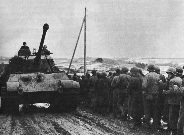 A Tiger II of schwere SS Panzer Abteilung 501 advances west past a column of American prisoners of the 99th Infantry Division captured at Honsfeld and Lanzerath.