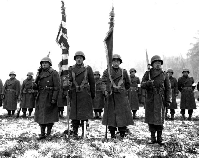 The 100th Eventually merged with the 442nd Infantry Battalion. Here, two color guards and color bearers of the Japanese-American 442d Combat Team, stand at attention, while their citations are read. They are standing on ground in the Bruyeres area, France, where many of their comrades fell. Photo - Wikipedia / Public Domain