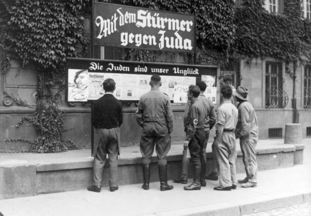 German citizens, publicly reading Der Stürmer, in Worms, 1933. The billboard heading reads: "With the Stürmer against Judea"; By Bundesarchiv, Bild 133-075 / CC-BY-SA 3.0, CC BY-SA 3.0 de, https://commons.wikimedia.org/w/index.php?curid=5338071 