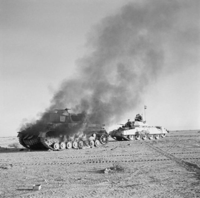 British Crusader tank passes a buning German PzKpfw IV; Photo Source