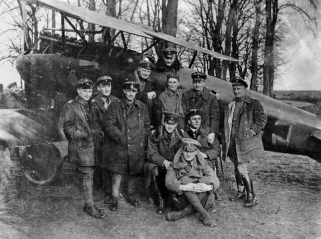 Manfred von Richthofen (in the cockpit) by his famous Rote Flugzeug (“Red Aircraft”), with other members of Jasta 11. His brother, Lothar, is seated on the ground. Photographed 23 April 1917. By Bundesarchiv – CC BY-SA 3.0 de