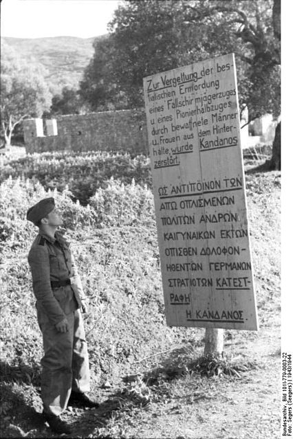 A German soldier standing in front of a sign in Greece that states: In retaliation for the brutal murder of paratroopers and combat engineers in an ambush by armed men and women, Kandanos was destroyed. By By Bundesarchiv – CC BY-SA 3.0 de