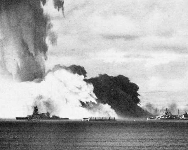 As the spray column falls, a radioactive “base surge,” like mist at the bottom of a waterfall, moves out toward the target ships. Foreground ship (left) is the 725 feet (221 m) long Japanese battleship Nagato.