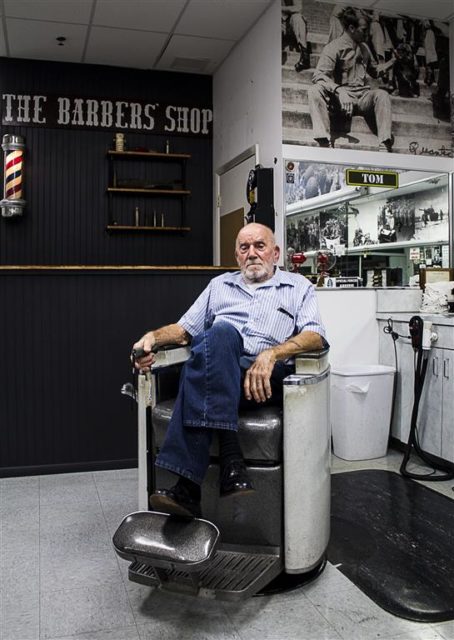 Tom Sakezles, a WWII veteran, and owner of The Barbers’ Shop in Tampa sits in the chair where he used to cut hair. Above him in the upper right of the photo is a picture of him from his military service. Photo by James Judge.
