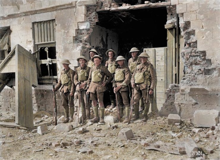 A group of eight British soldiers stand next to a blown-out building. Photo colourised by Royston Leonard / mediadrumworld.com