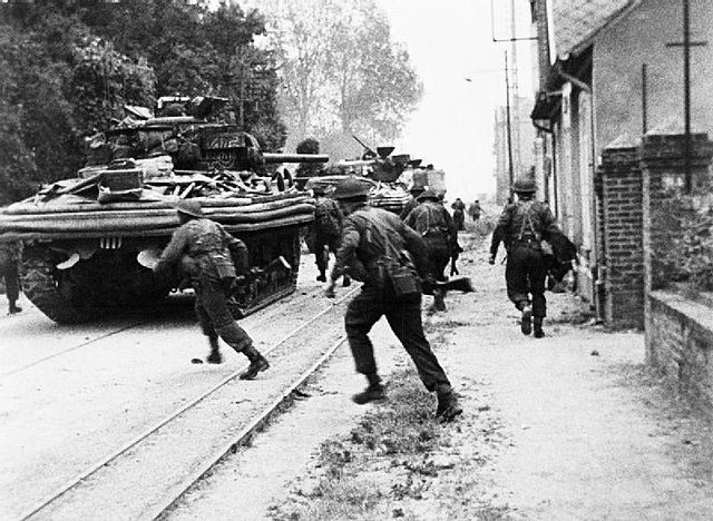 Men of No 4 Commando engaged in house to house fighting with the Germans at Riva Bella, near Ouistreham. Sherman DD tanks of ‘B’ Squadron, 13/18th Royal Hussars are providing fire support and cover. After subduing the opposition, No 4 Commando moved inland to link up with 6th Airborne Division.