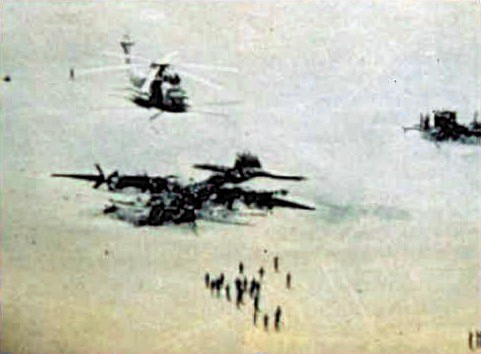 A photo of the “Desert One” landing site, a piece of desert in Iran used by U.S. forces as a refueling point in an attempt to rescue U.S. hostages in Iran. On 24 April 1980 a U.S. Navy Sikorsky RH-53D Sea Stallion (BuNo 158760, visible at right) collided with a U.S. Air Force Lockheed EC-130E Hercules (s/n 62-1809, wrecked in the foreground) during refueling after the mission was aborted. Both aircraft were destroyed, eight crewmen died. In the background is one of the five intact, but abandoned RH-53Ds. Original caption: “Wreckage at Desert One, Iran (April 1980) where eight Americans died.”