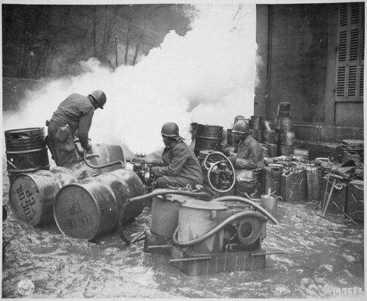 Soldiers of the 161st Chemical Smoke Generating Company, U.S. Third Army, move a barrel of oil in preparation to refilling an M-2 smoke generator, which spews forth a heavy cloud of white smoke. These men are engaged in laying a smoke screen to cover bridge building activities across the Saar River near Wallerfangen, Germany, December 1944
