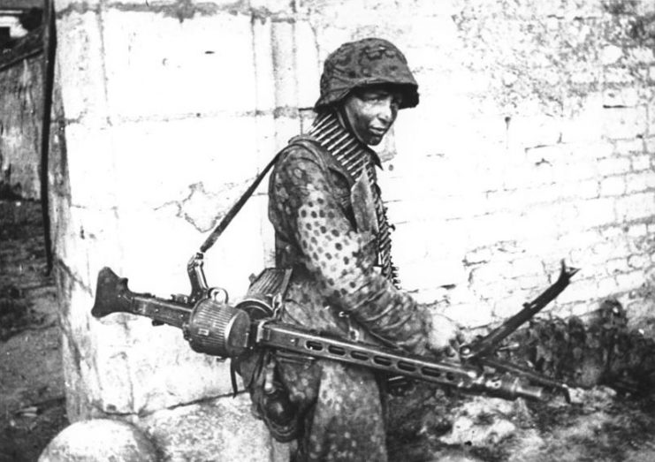 A German Waffen SS soldier involved in heavy fighting in and around the French town of Caen in mid-1944. He is carrying an MG 42 configured as a light support weapon with a folding bipod and detachable 50-round belt drum container. Photo: Bundesarchiv, Bild 146-1983-109-14A / Woscidlo, Wilfried / CC-BY-SA 3.0