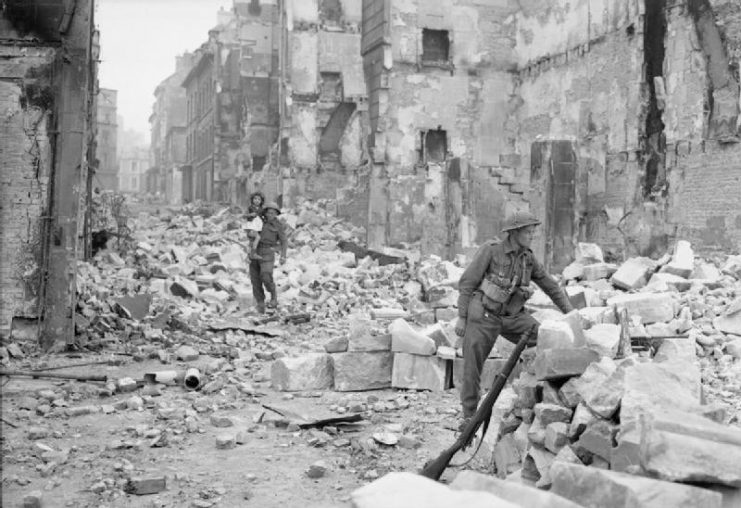 A British soldier in Caen after its liberation, gives a helping hand to an old lady amongst the scene of utter devastation. The town had been substantially destroyed during the bombardment, leaving large numbers of the population homeless.