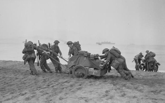 Troops making their way inland after landing at Algiers. Here some men are pulling and pushing a trailer of equipment over the sandy beach, and in the background two soldiers are manhandling a motorbike.