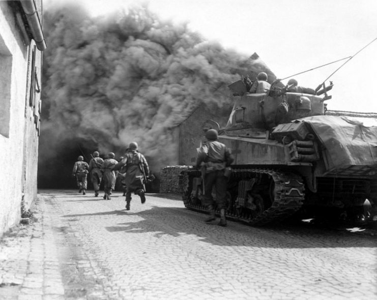 US Army soldiers supported by a M4 Sherman tank move through a smoke filled street in Wernberg, Germany during April 1945