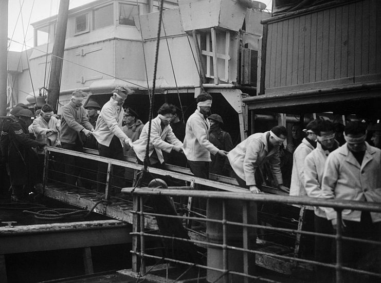 Blindfolded SCHARNHORST survivors, in merchant seaman rescue kit, walking down a gang-plank on their way to internment.