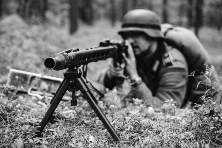 Gomel, Belarus – June 20, 2014: Hidden Unidentified Re-enactor Dressed As German Wehrmacht Soldier Aiming A Machine Gun At Enemy From Trench In Forest.
