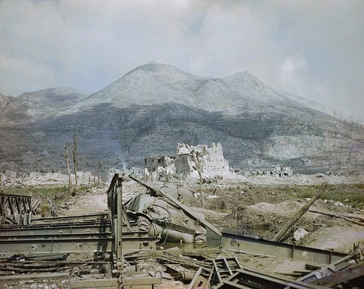 View of Cassino after heavy bombardment showing a knocked out Sherman tank by a Bailey bridge in the foreground, with Monastery Ridge and Castle Hill in the background.