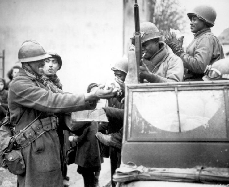 Two smiling French soldiers fill the hands of American soldiers with candy, in Rouffach, France.