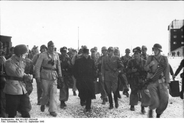 Mussolini, surrounded by German and Italian officers and soldiers, heads for the plane.