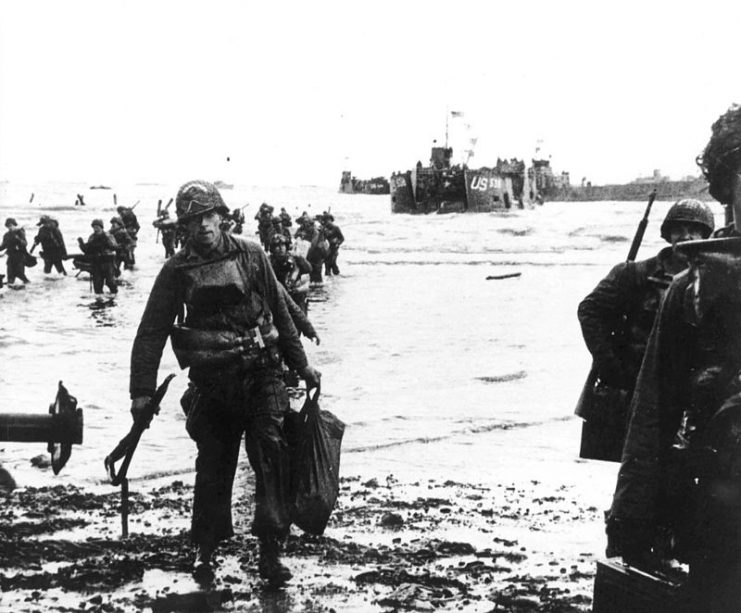 Carrying their equipment, U.S. assault troops move onto Utah Beach. Landing craft can be seen in the background.