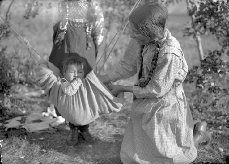 Cree Girl Playing With a Child Suspended in a Small Hammock – Waterhen River, Northern Saskatchewan