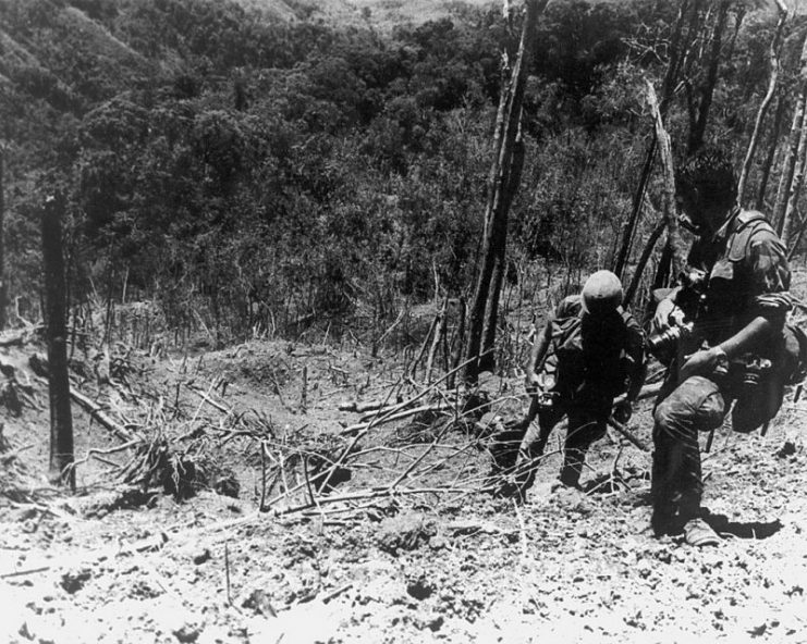 This image shows a U. S. Army Photographer and assistant climbing through the devastated landscape on Dong Ap Bia after the battle. (Melvin Zais Photograph Collection). (Photo Credit: USAMHI)
