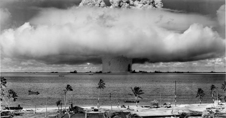 Mushroom-shaped cloud and water column from the underwater explosion Baker nuclear explosion of July 25, 1946. Photo taken from a tower on Bikini Island. The Wilson clouds lift, revealing the full spray column. The battleship Arkansas is right of column, along with other ships.