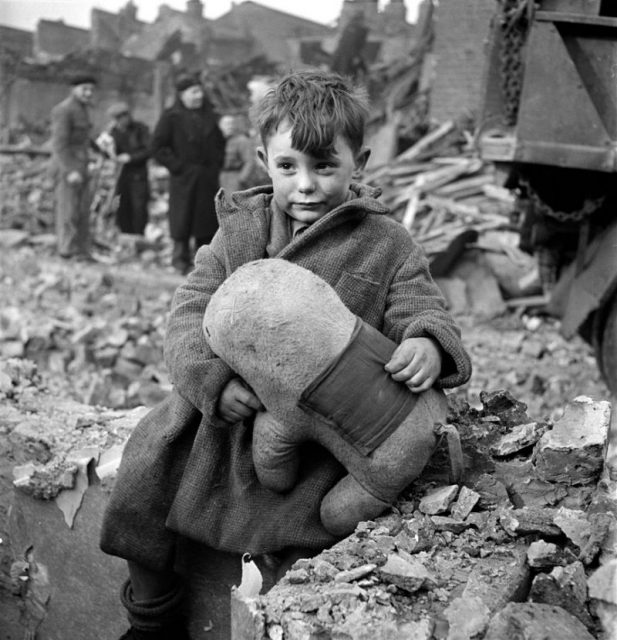 Abandoned boy holding a stuffed toy animal amid ruins following German aerial bombing of London, England. Date 1945