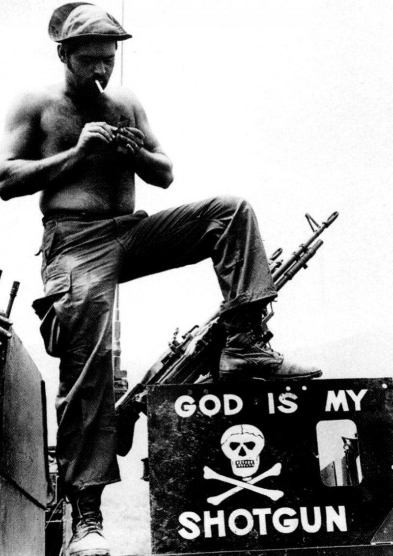 Khe Sanh, South Vietnam, April 12, 1971 - An American soldier, lighting a cigarette in front of his machine gun atop a vehicle, stands above a sign serving as testament to his battlefield beliefs.