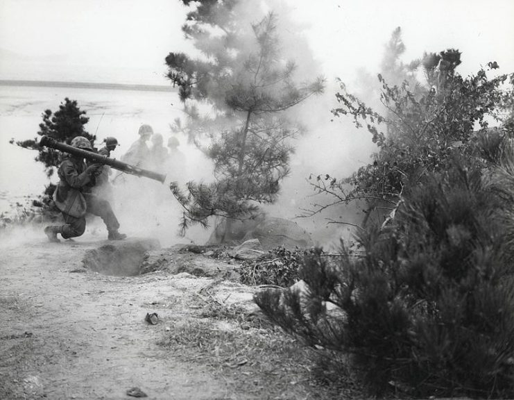 “Marine riflemen in background stand by while their 3.5 bazooka man puts a round into a Communist position down the hill. This action took place in mopping-up operations in Korea.