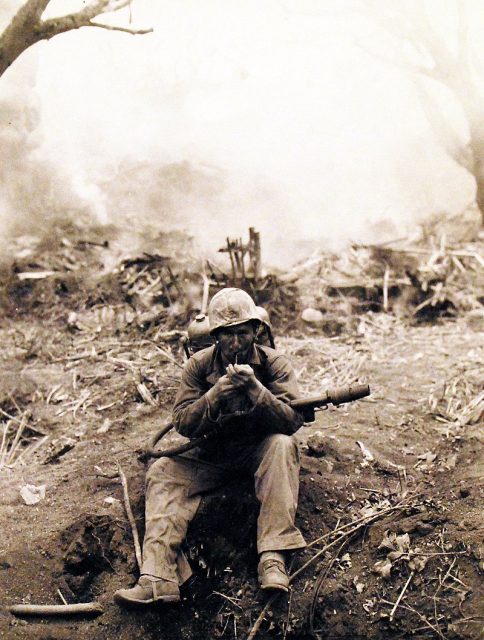Private First Class Wilfred Voegeli armed with a flame thrower, halts to light up his pipe on Iwo Jima