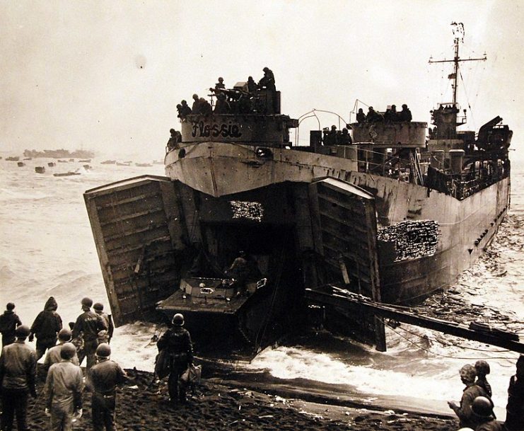 Flat-nose Flossie” LST at beach while elements of a Marine Corps amphibious tractor unit unload her cargo under protection of her forward guns, February 19, 1945.