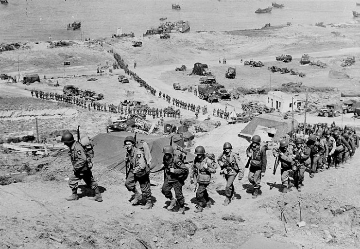 2nd Infantry Division marching up the bluff at the E-1 draw in the Easy Red sector of Omaha Beach on D+1, 7 June 1944. They are going past the German bunker, Widerstandsnest 65, that defended the route up the Ruquet Valley to Saint-Laurent-sur-Mer.