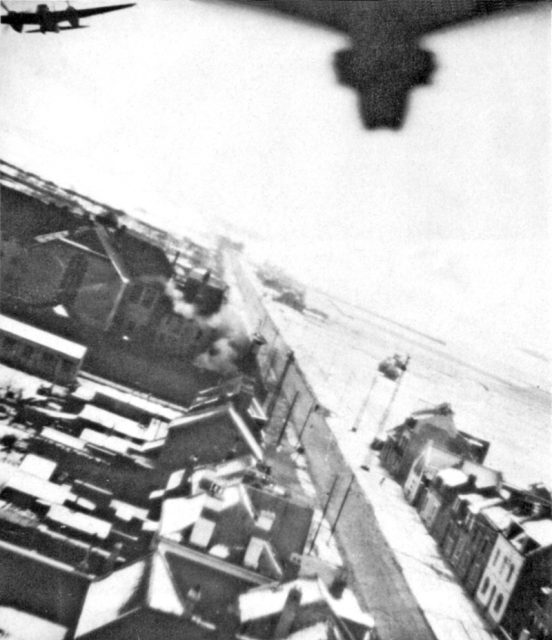Operation Jericho – Amiens Prison during the raid – picture taken from the accompanying PRU Mosquito (the fuselage & tailwheel of which appears in the top-right of the picture) and also showing one of the attacking Mosquitoes (with bomb-doors open) at the extreme top-left of the picture. The Prison itself is the large, dark building, at the center-left.