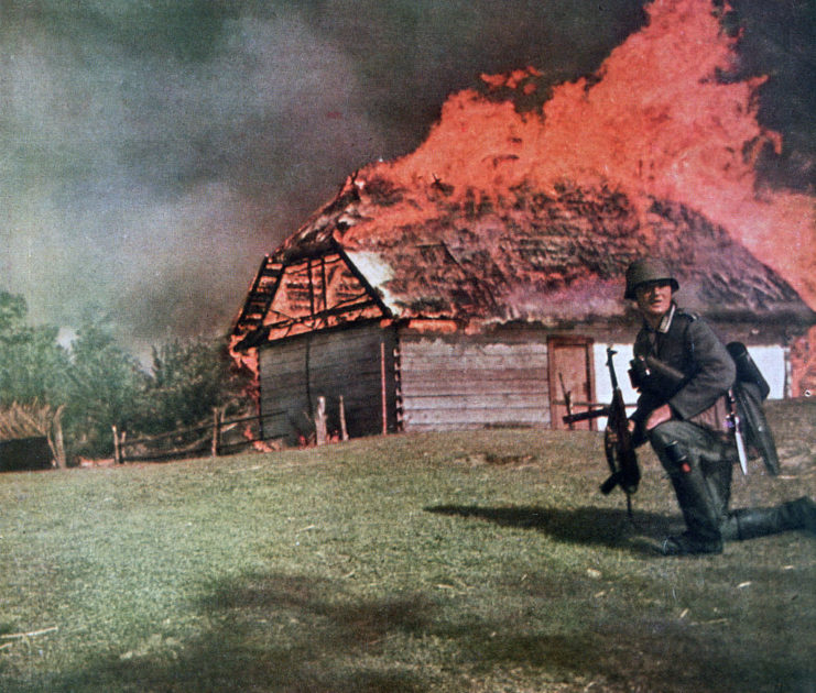 A German soldier kneeling in front of a burning building with his rifle in his hand