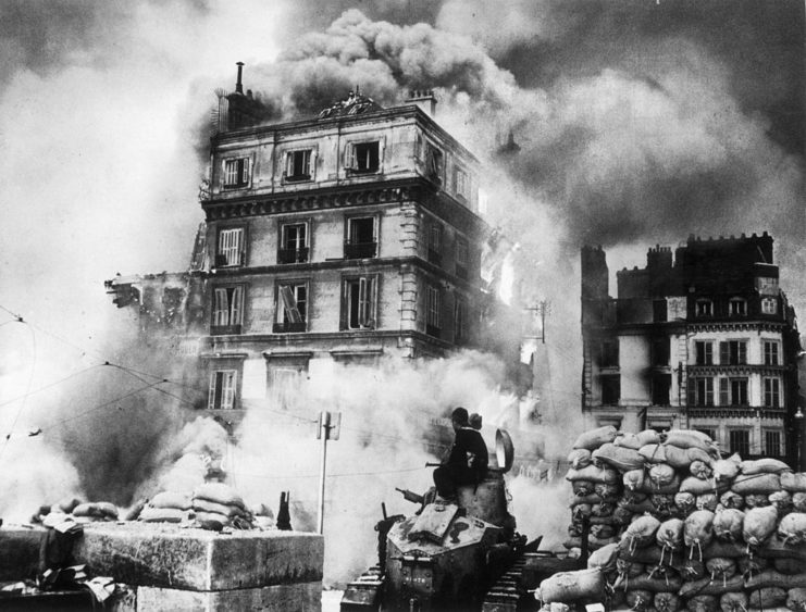 A tall building on fire, surrounded by sandbags, while two soldiers travel away on a military vehicle