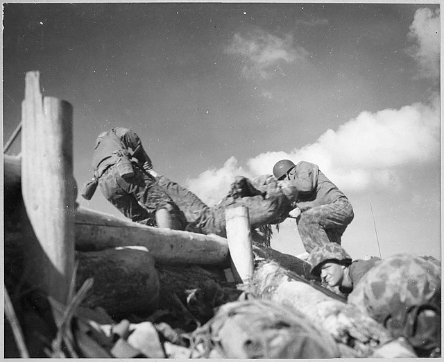 Two Marines carry another Marine over a wooden fence