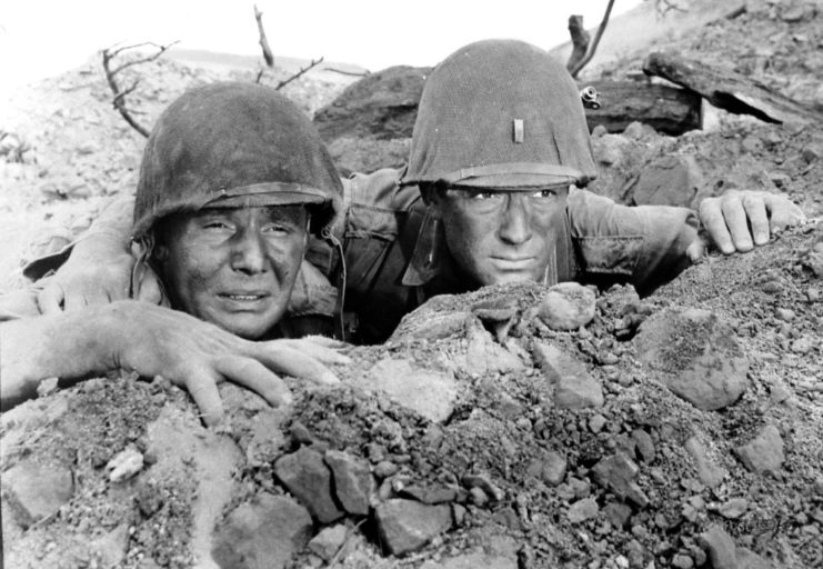 Two men wearing helmets, looking out of a trench